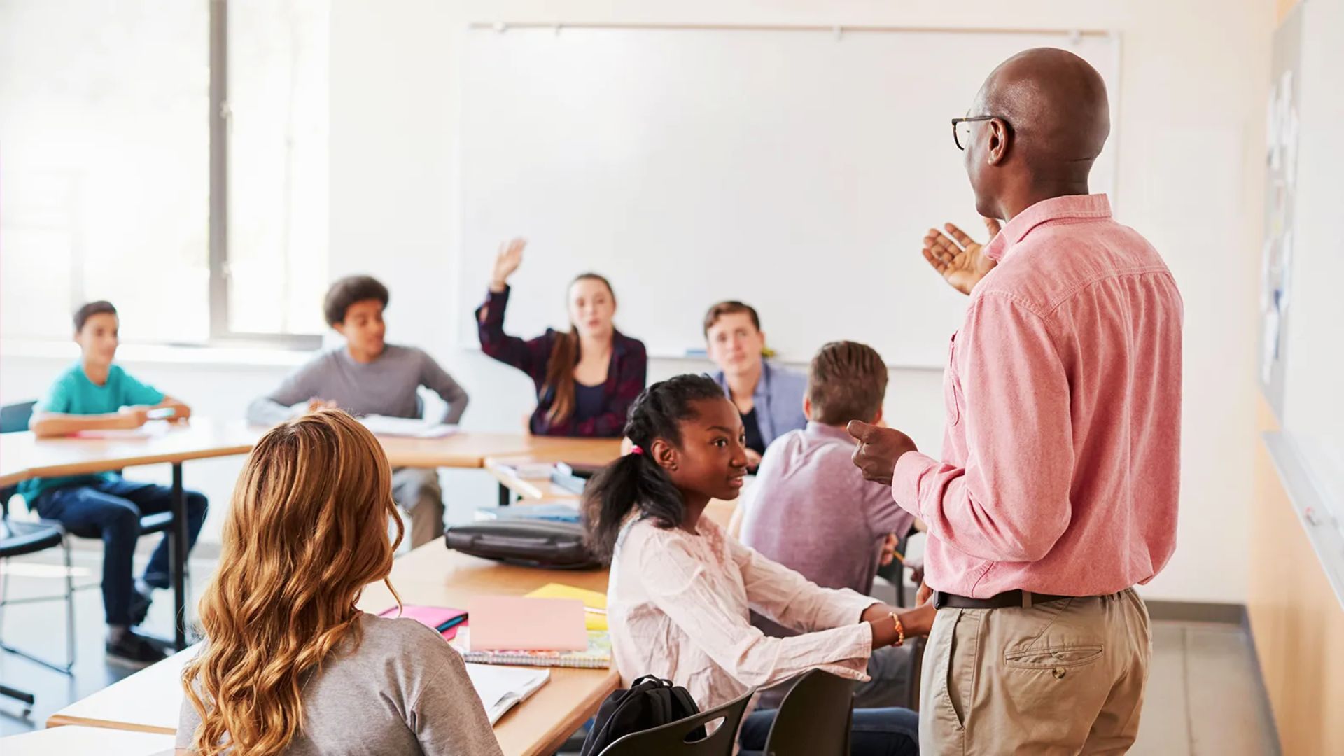 A teacher leads a discussion while students listen and one raises a hand to participate.