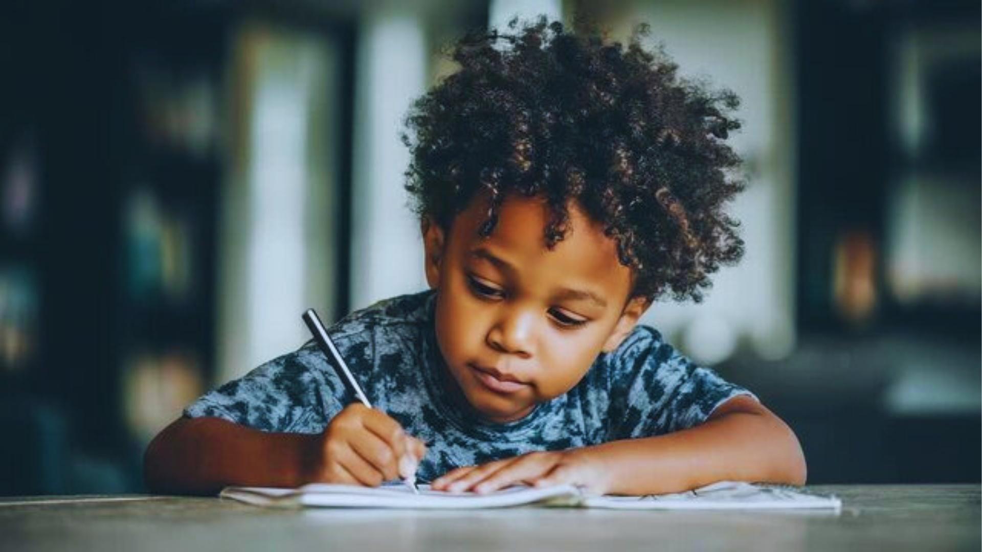 Focused boy with curly hair writing in notebook with black pen at wooden table in library.