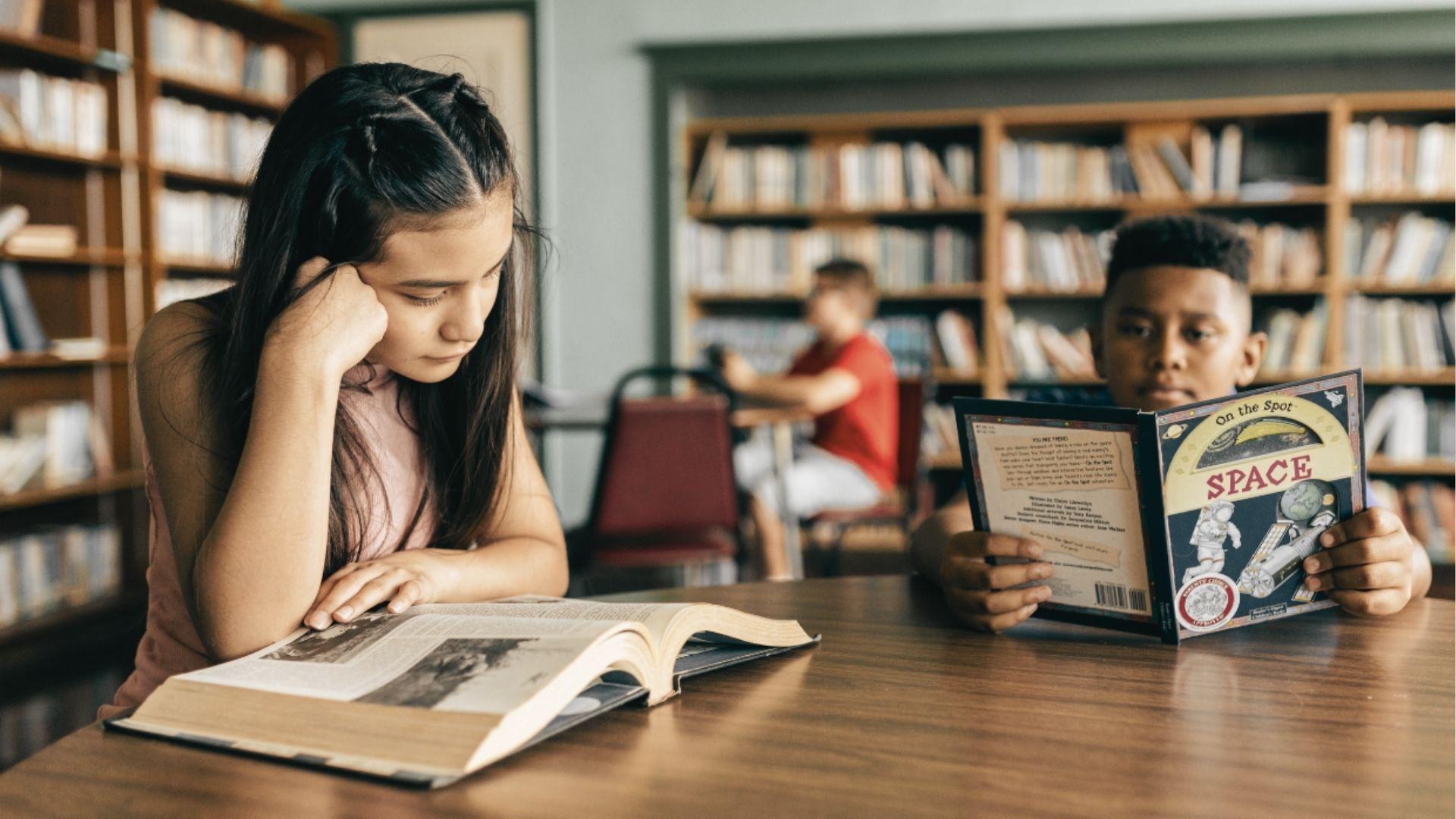 Two students read different books at a library table, one focused on an open textbook and the other on a space book.