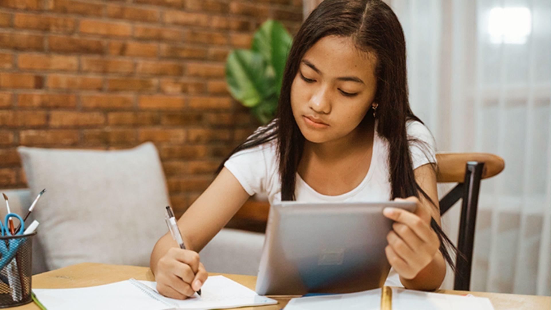 Girl taking notes from tablet while studying at home desk to practice balanced tech and writing.