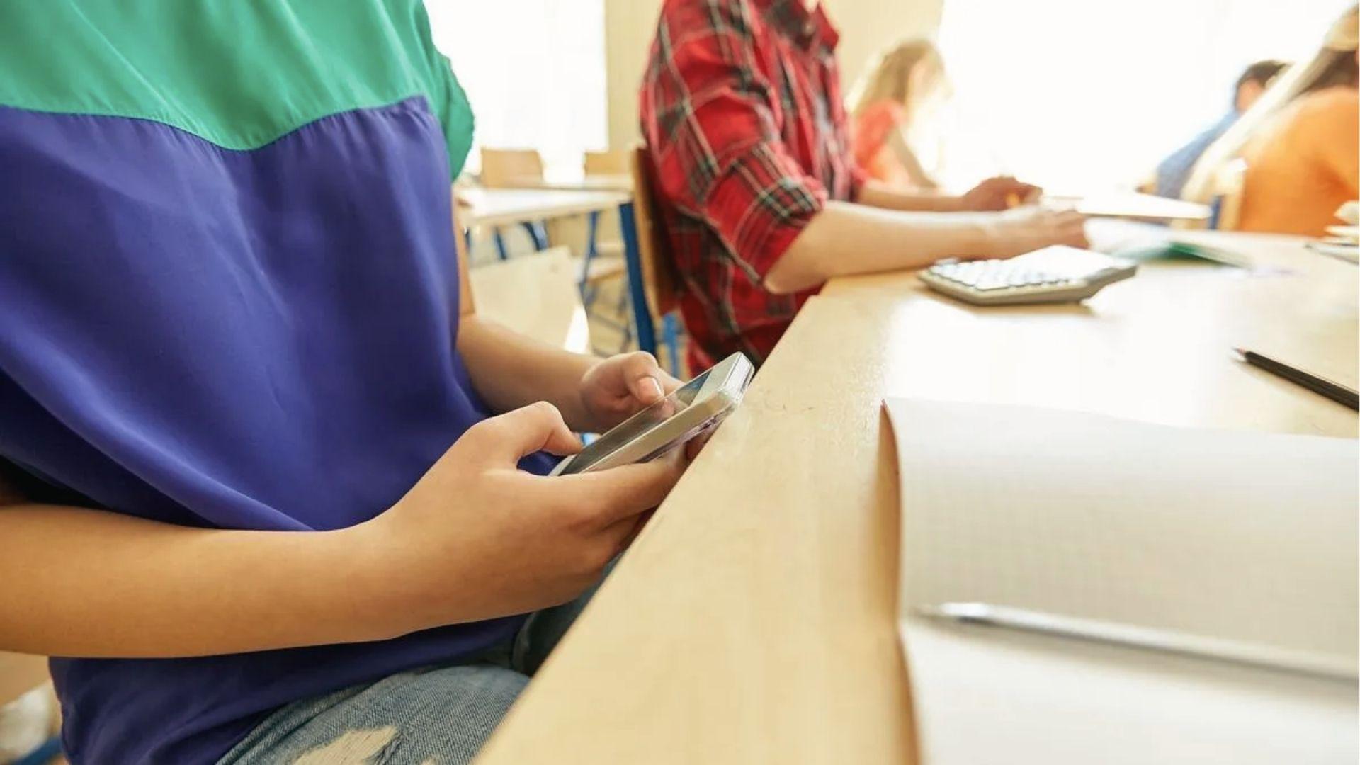 Student hides phone under desk in class, ignoring homework to scroll and procrastinate during study time.