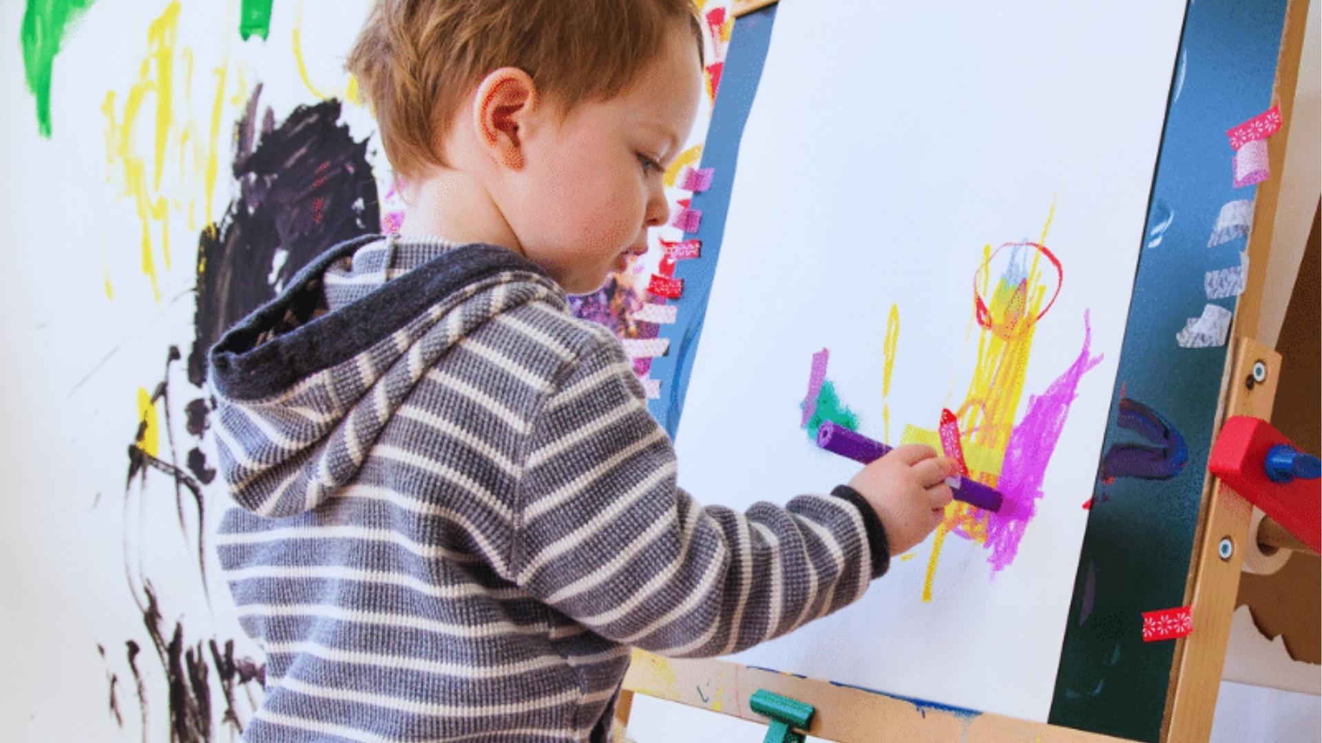 Young child in striped hoodie painting with purple marker on easel paper decorated with colorful washi tape.