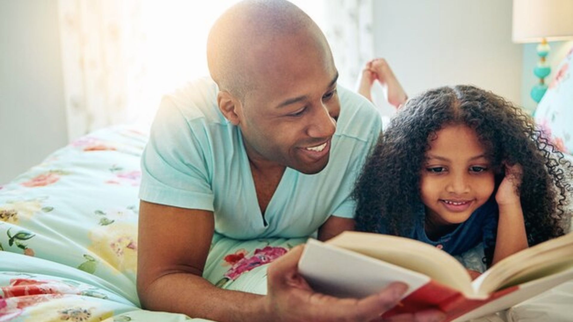 A father and daughter lie on a colorful bed, reading a book together with warm smiles.