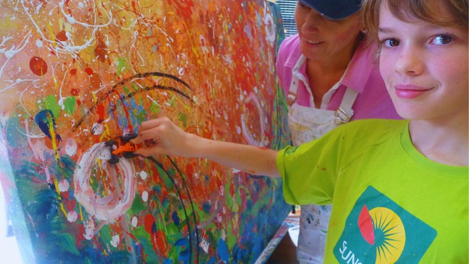 Boy in lime green shirt using toy car to paint colorful abstract splatters on large canvas with teacher.