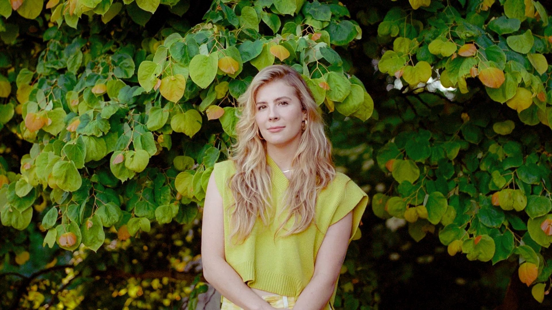 A woman in a yellow dress stands confidently in front of a large, leafy tree.