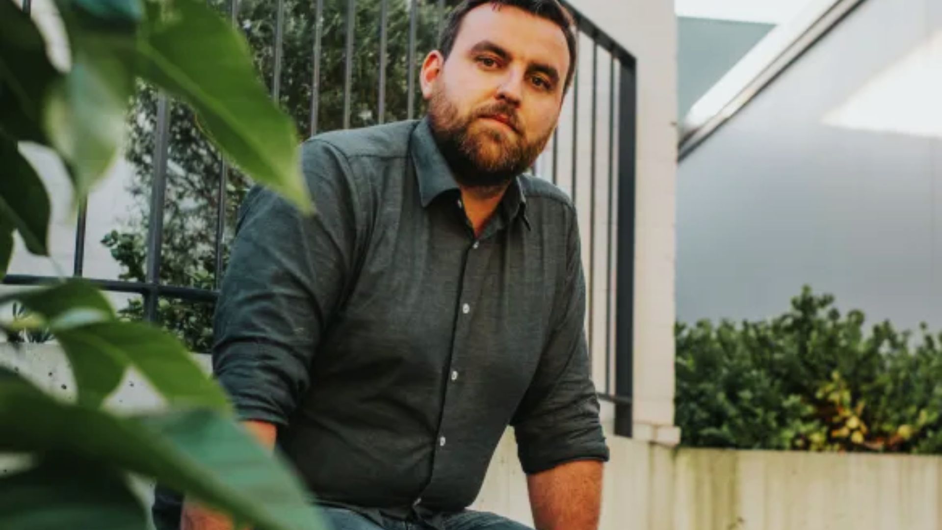 Benjamin Stevenson, an Australian author, sits on a ledge, wearing a shirt and sporting a beard.