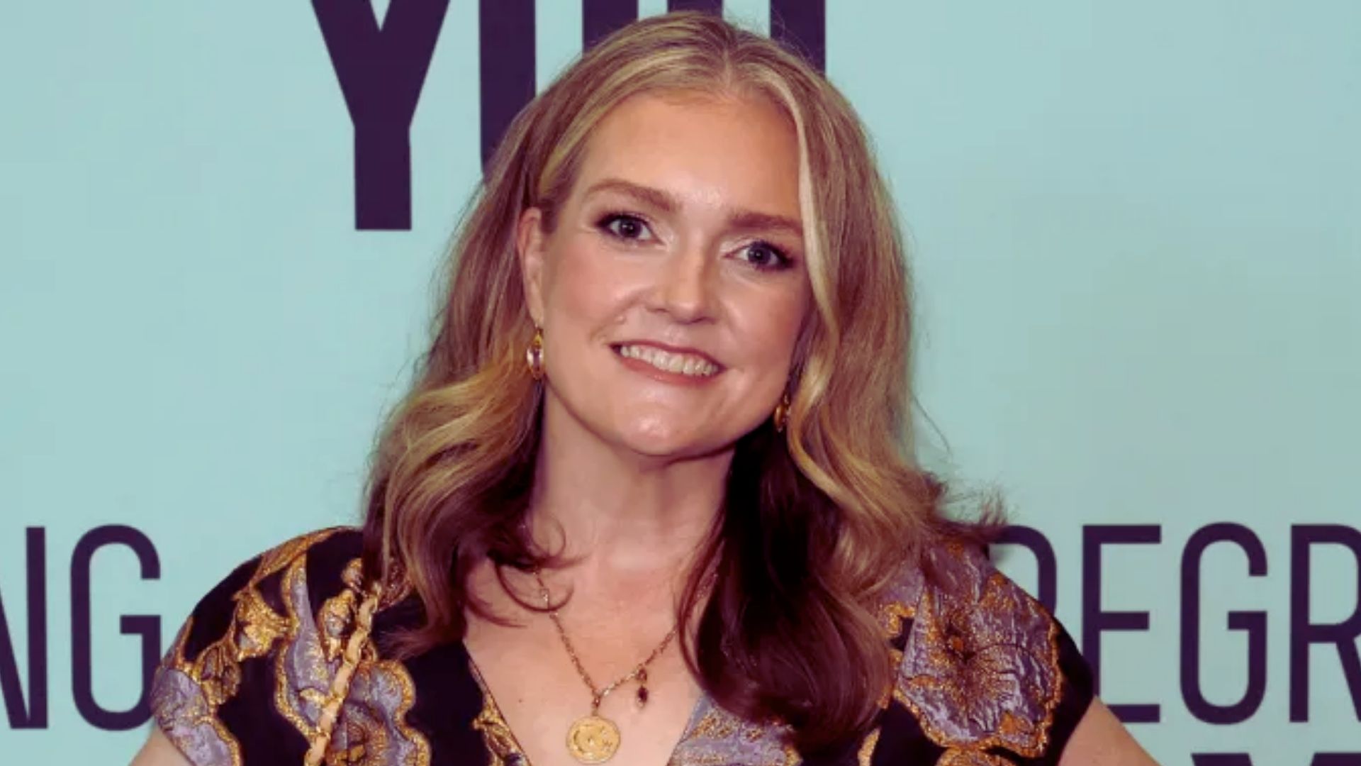 A woman with long hair wearing a gold necklace, looking confidently at the camera.