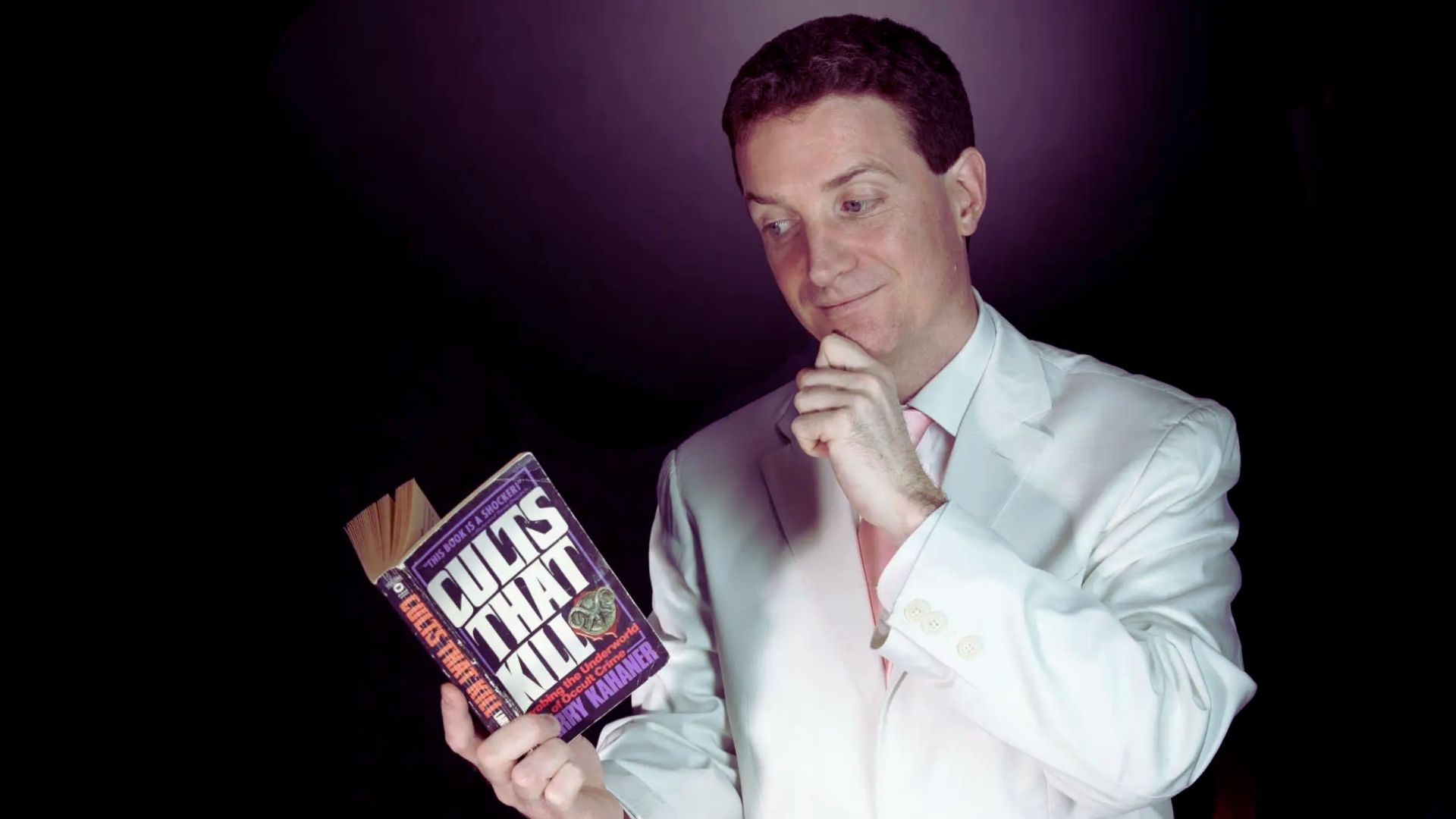 A man in a white suit stands holding a book, exuding a sophisticated and polished appearance.