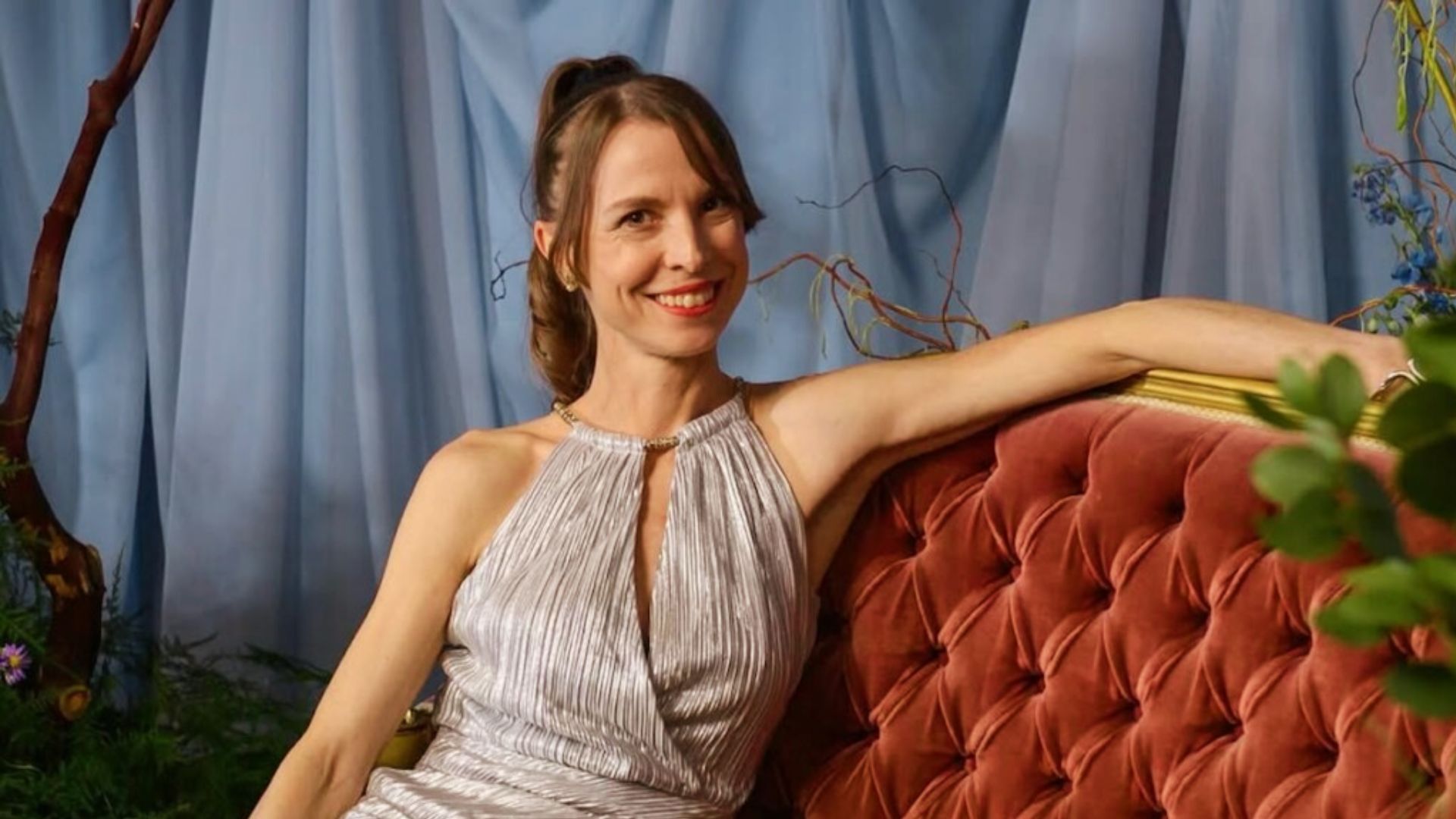 Nita Prose, Canadian author of "The Maid," sits on a red couch holding a flower in her hand.