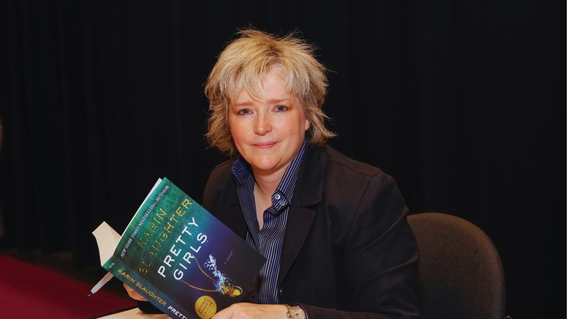 Author Karin Slaughter in black blazer and striped shirt smiles while holding Pretty Girls advance reader copy at book signing event.