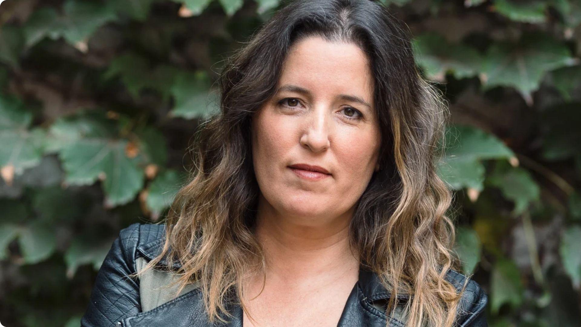 Agustina with dark wavy hair in a leather jacket posing in front of ivy-covered wall.