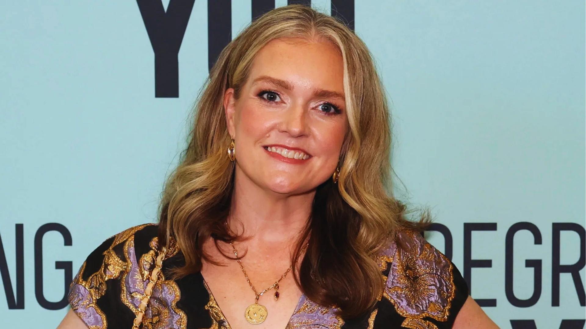 Author Colleen Hoover smiles at a media event wearing an ornate black and purple dress with gold jewelry against a light blue backdrop.