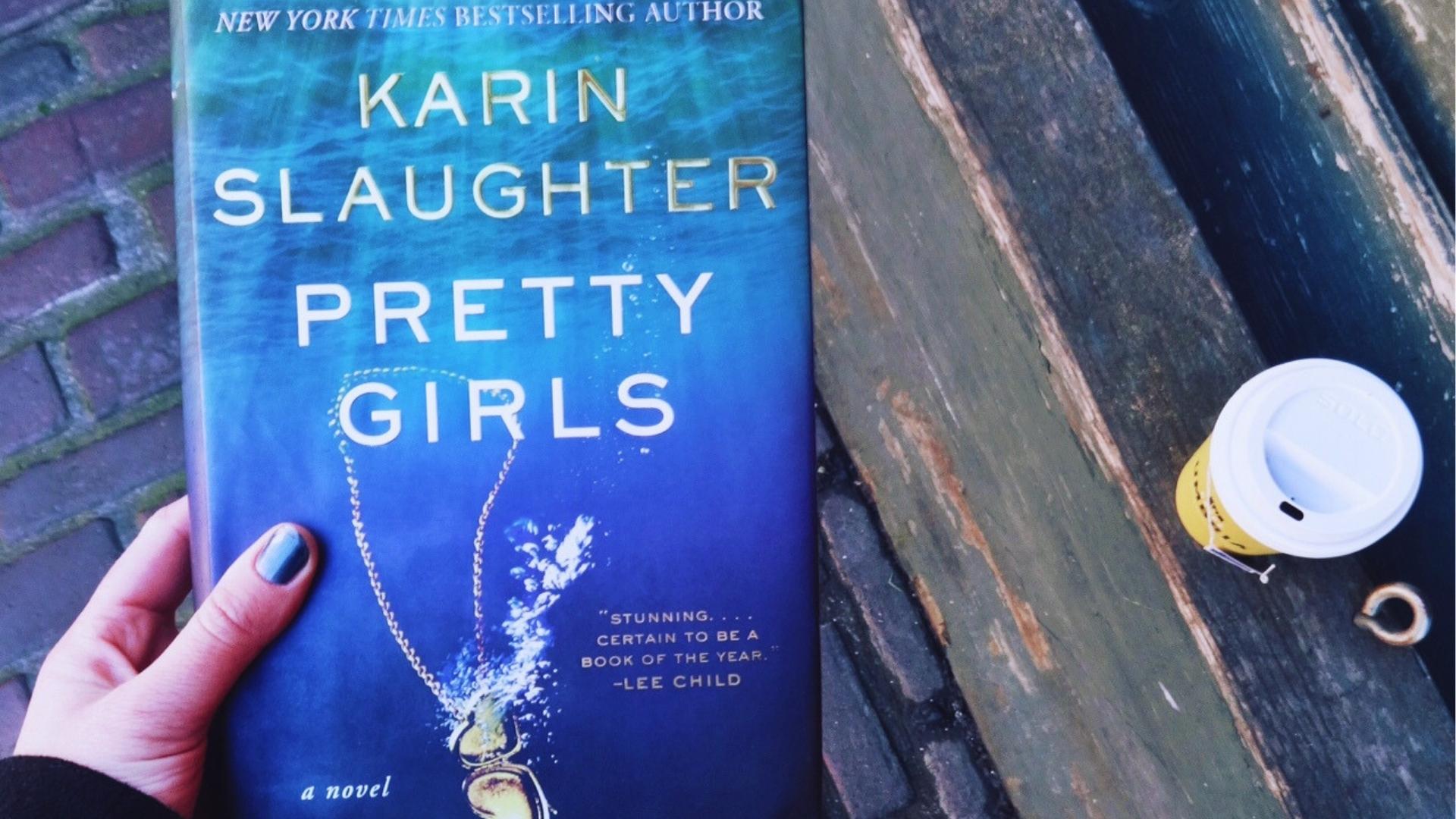 Hand with metallic blue nail polish holds Pretty Girls by Karin Slaughter hardcover beside yellow coffee cup on weathered bench.