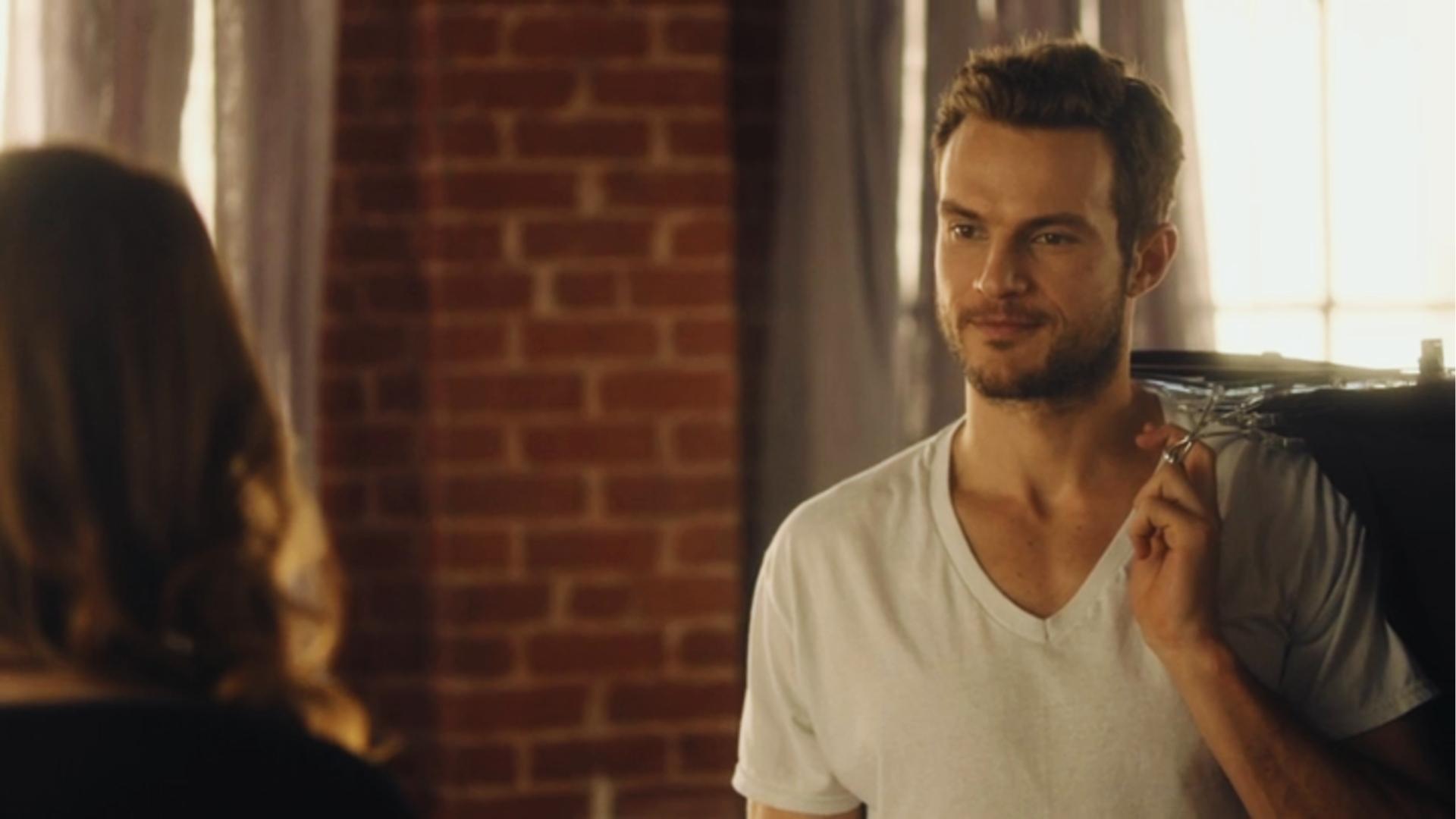 Man in white V-neck shirt holds jacket over shoulder while talking to woman, brick wall and windows in sunlit loft.
