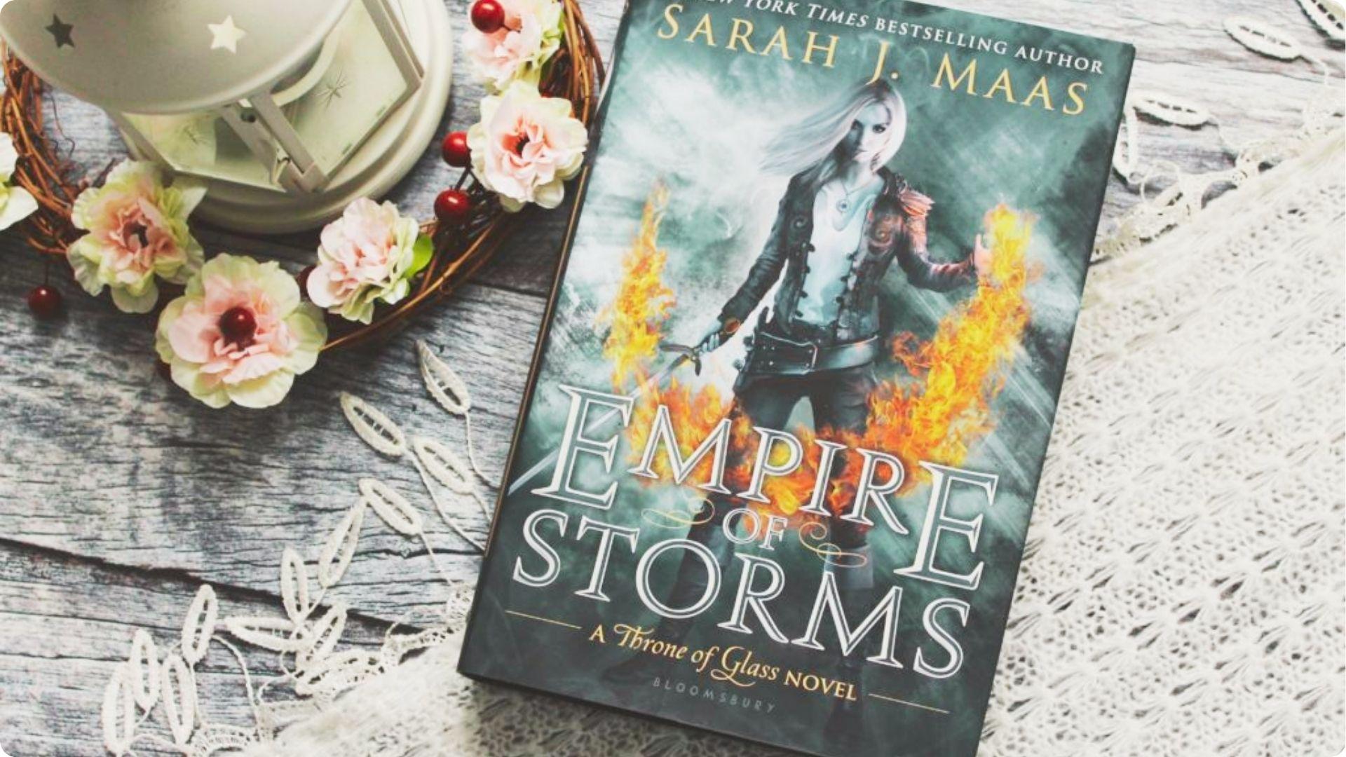 Empire of Storms by Sarah J. Maas lies on a rustic wooden table surrounded by pink flowers and a lantern.