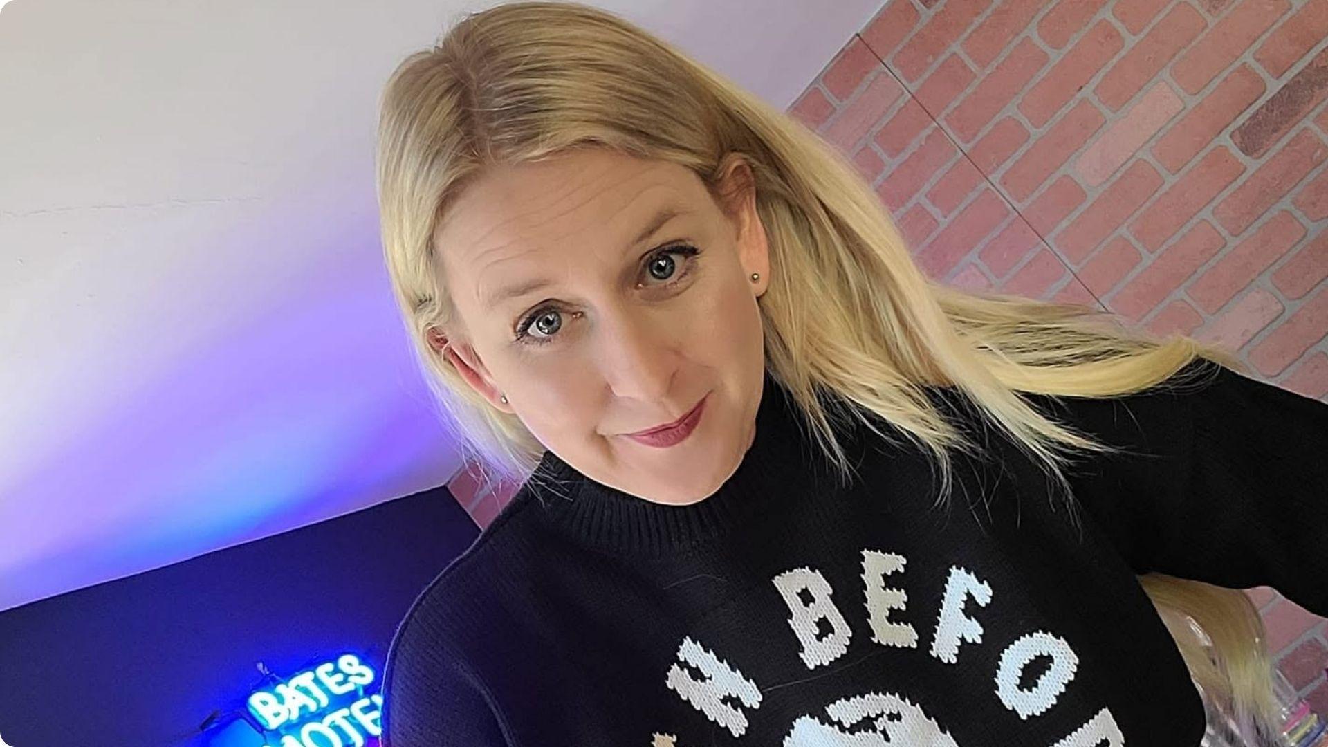Penelope Douglas in a black novelty sweater taking a selfie near a Bates Motel neon sign.