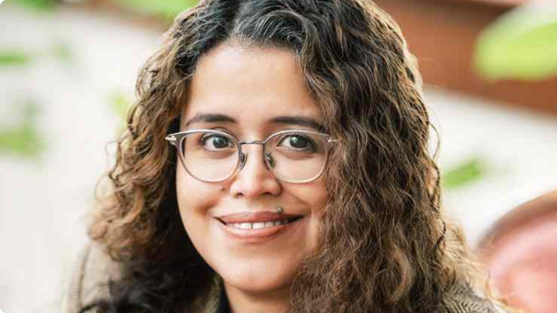 The author with curly hair and round glasses posing outdoors with greenery in the background.
