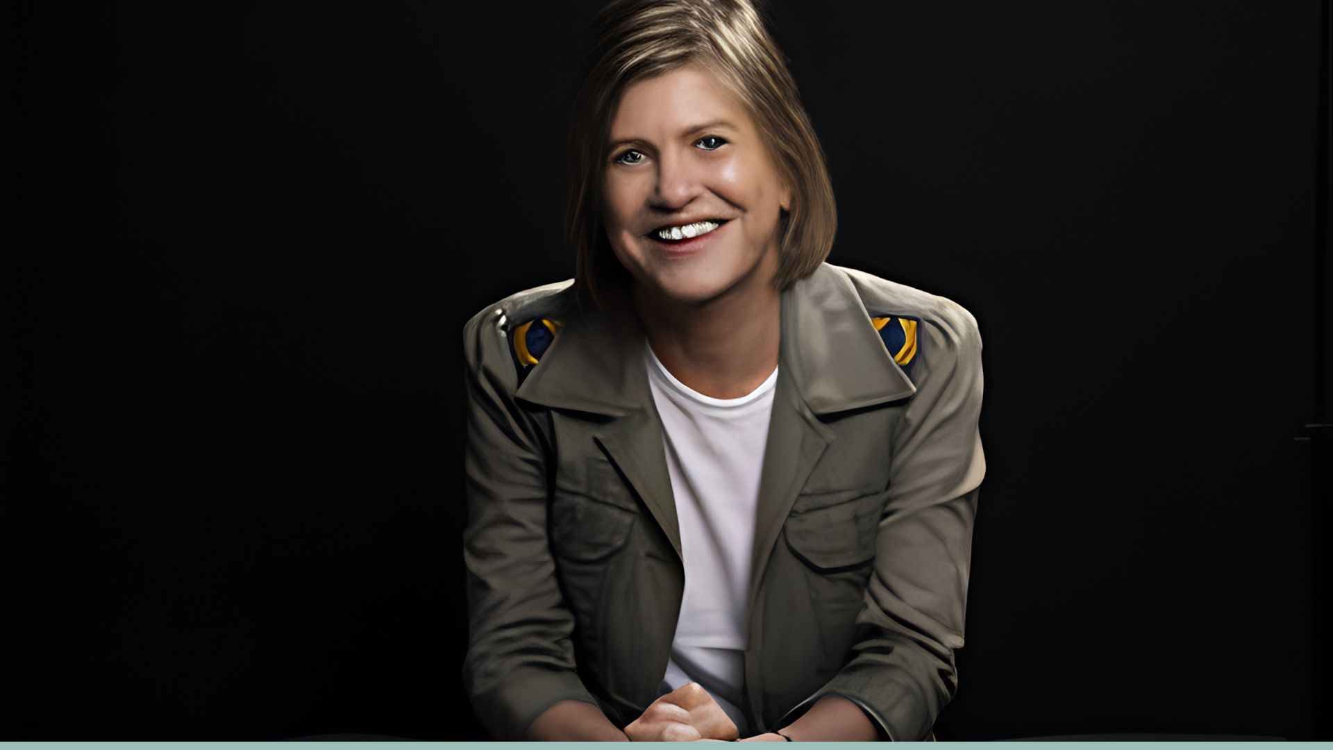 Author Clare Leslie Hall wearing olive jacket and white shirt, smiling against black background in studio portrait.