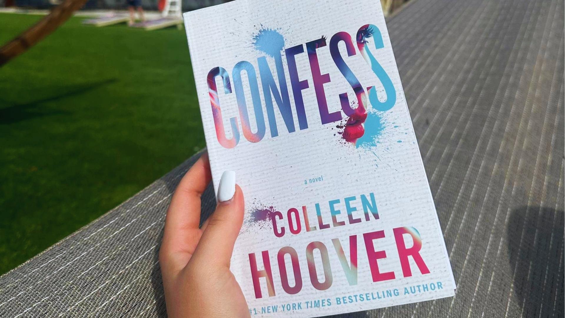Hand with white nail polish holds Confess by Colleen Hoover paperback featuring blue watercolor cover and sinking locket near outdoor lounge.