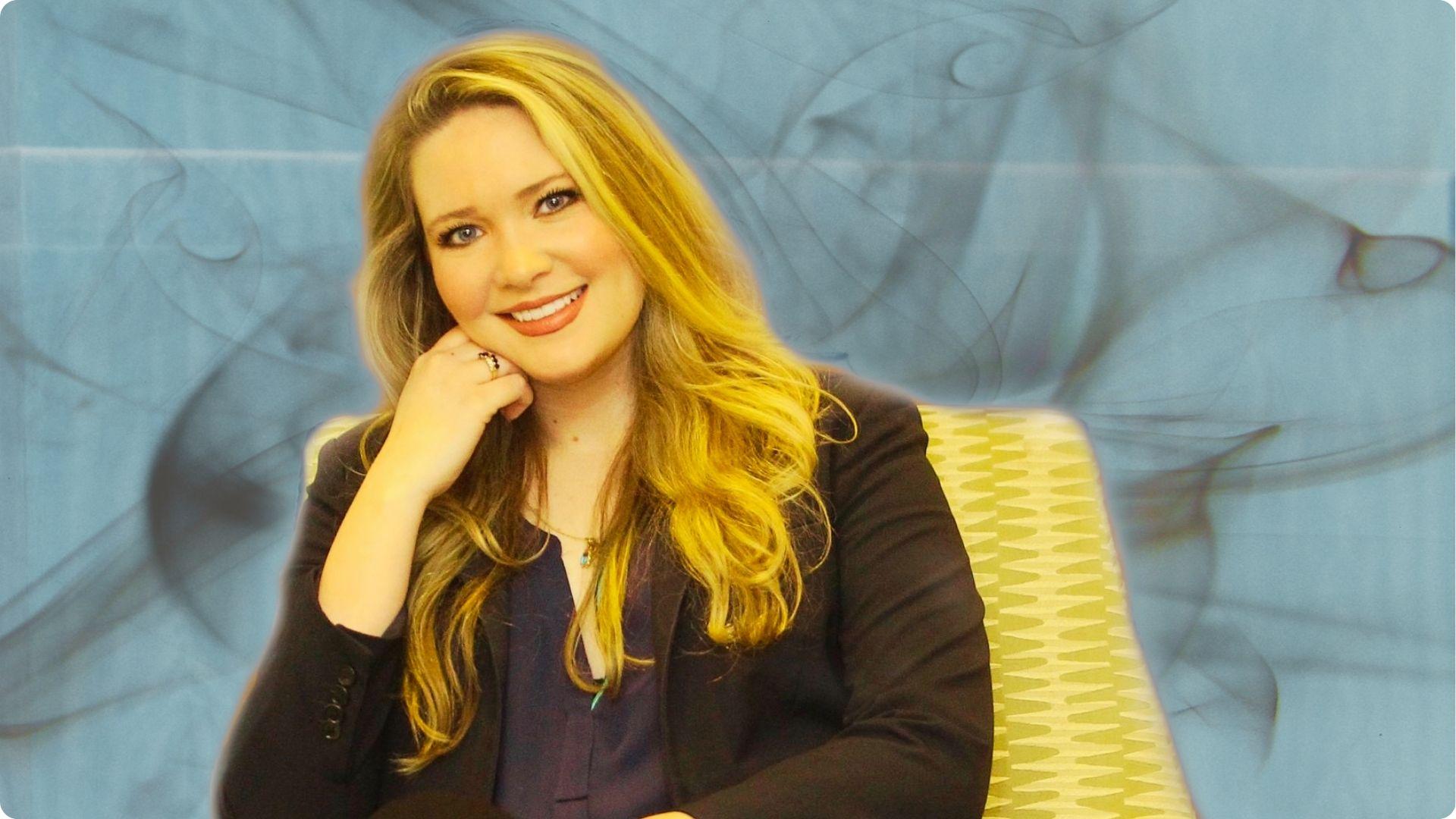 Sarah J. Maas Smiling blonde in a black blazer and navy top seated against an abstract blue background.