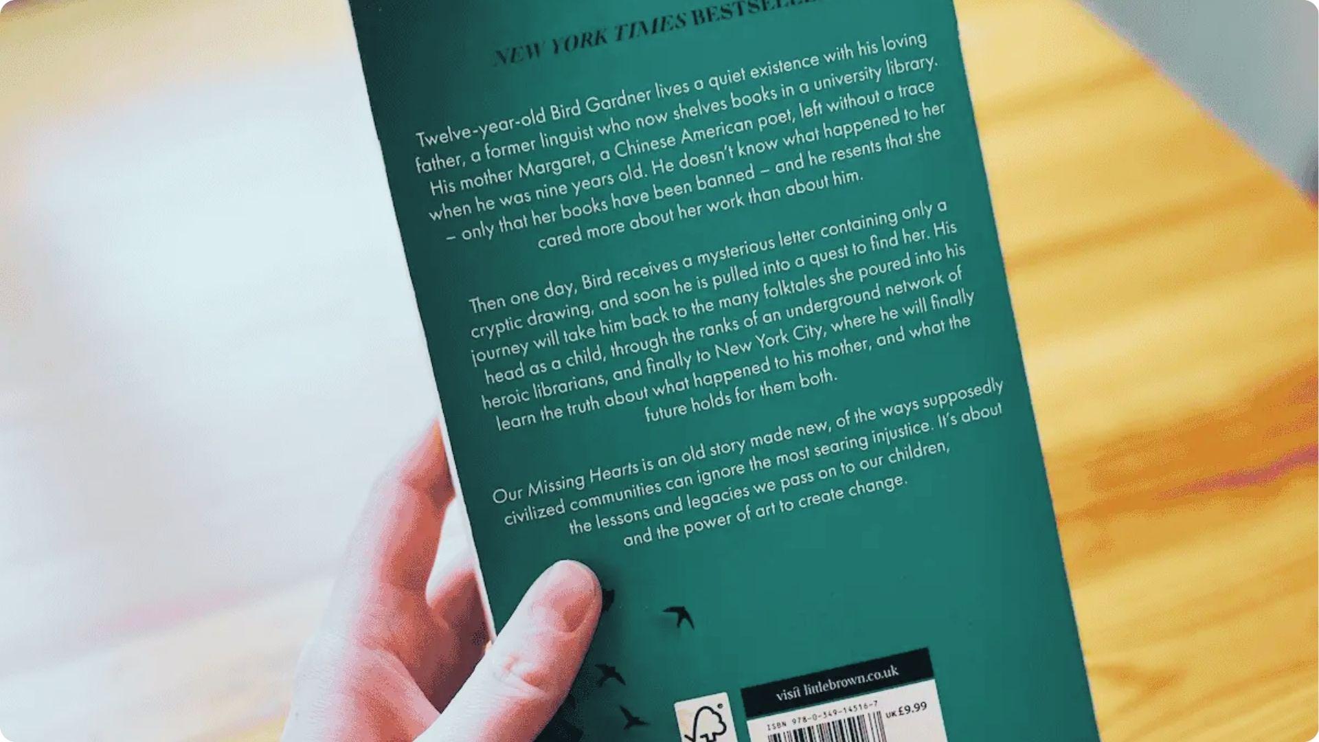 Hand holding a teal paperback showing the full back cover text for Our Missing Hearts.