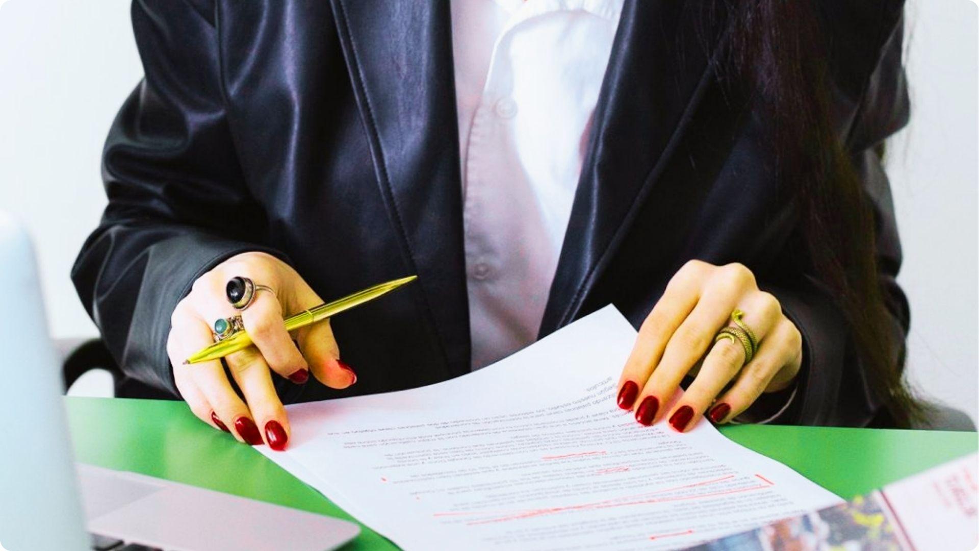 Close-up of hands with red nails and rings holding a gold pen over a document with red edits.