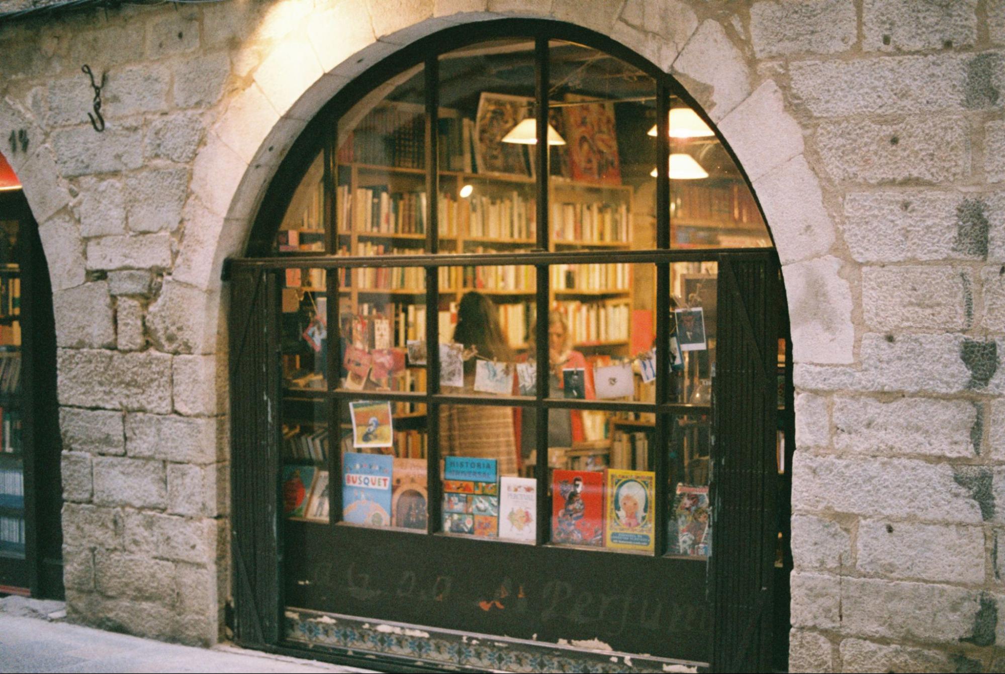 Bookshop with a large arched window in a stone façade, displaying books with shelves visible inside.