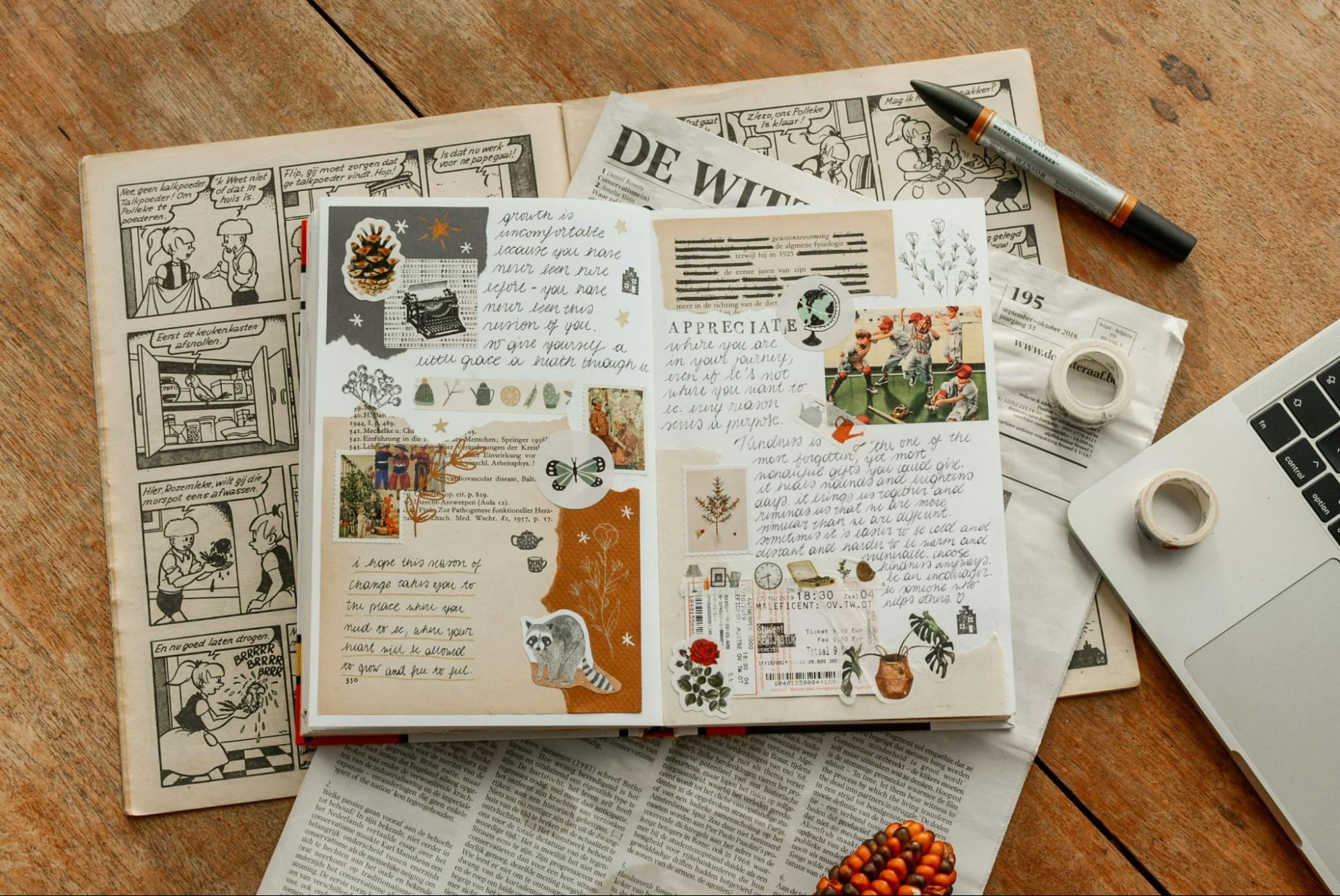 Open journal decorated with stickers, handwritten notes, and paper cutouts on a wooden desk, surrounded by newspapers and stationery.